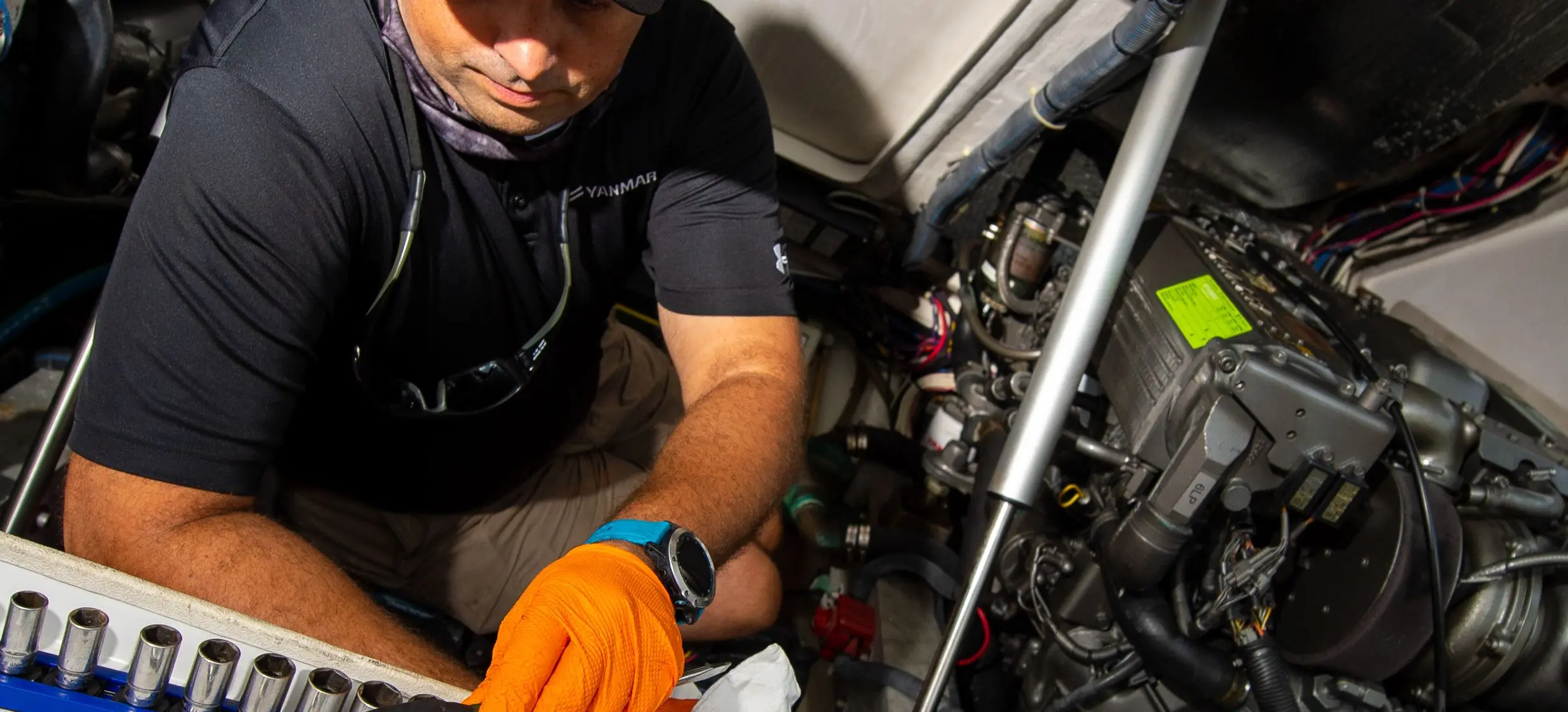 A YANMAR technician kneeling at an open engine hatch on a boat, a socket rail laid out beside him and the diesel engine to his right